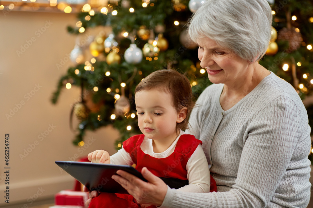 winter holidays and family concept - happy grandmother and baby granddaughter with tablet pc computer sitting near christmas tree at home winter holidays and family concept - happy grandmother and baby granddaughter with tablet pc computer sitting near christmas tree at home