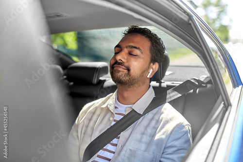 transportation, technology and people concept - happy smiling indian male passenger with wireless earphones on back seat of taxi car