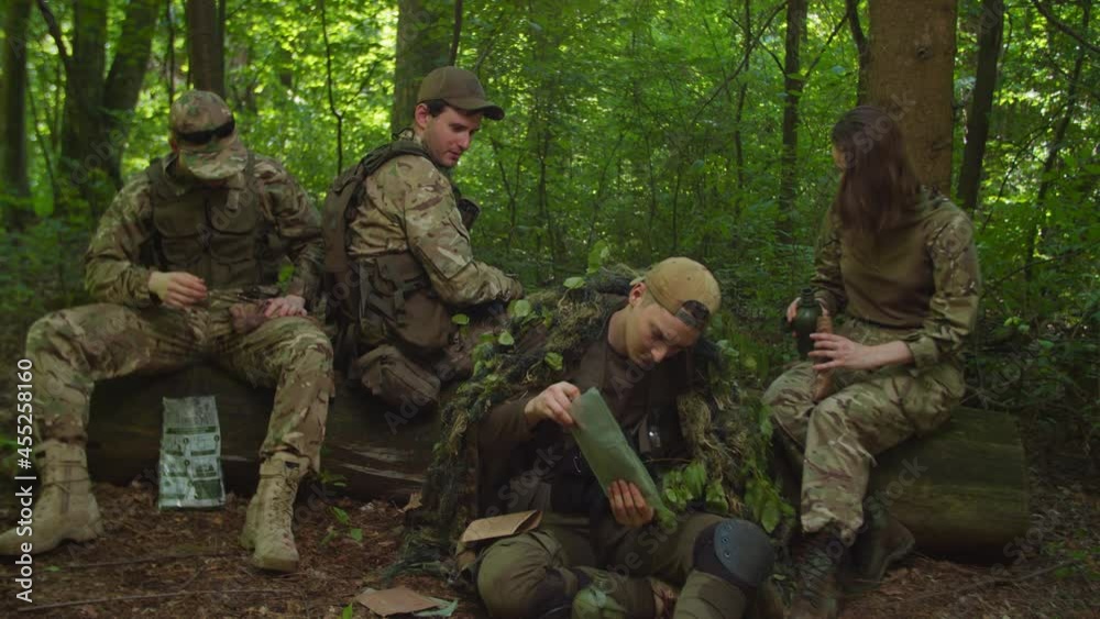 Group of military men and servicewoman in camouflage clothing resting in halt, preparing rations to eat in forest area. Soldiers smoking, relaxing during danger reconnaissance mission