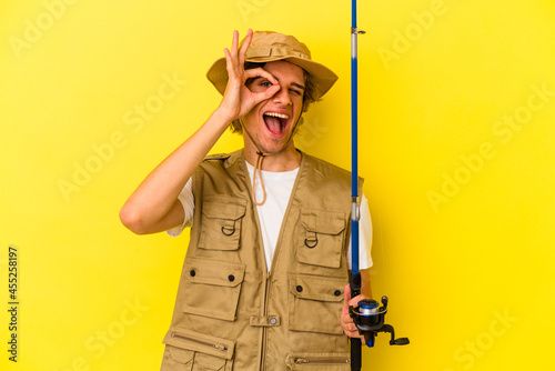 Young fisherman with makeup holding rod isolated on yellow background excited keeping ok gesture on eye.