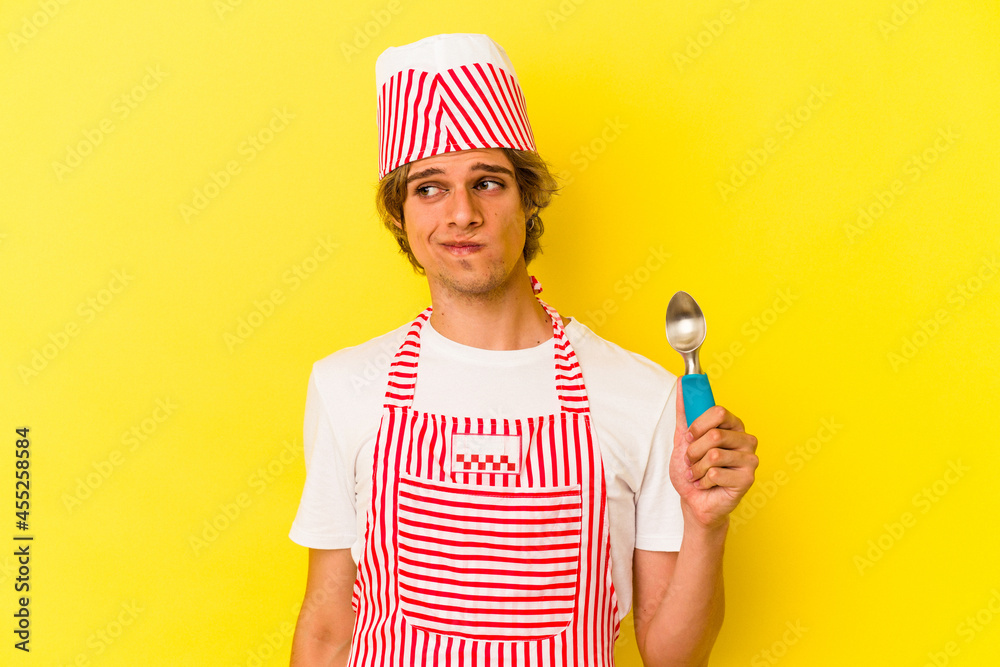 Young ice cream maker man with makeup holding spoon isolated on yellow background confused, feels doubtful and unsure. Young ice cream maker man with makeup holding spoon isolated on yellow background confused, feels doubtful and unsure.