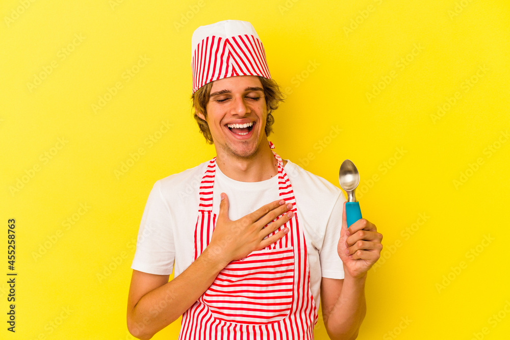 Young ice cream maker man with makeup holding spoon isolated on yellow background laughs out loudly keeping hand on chest. Young ice cream maker man with makeup holding spoon isolated on yellow background laughs out loudly keeping hand on chest.