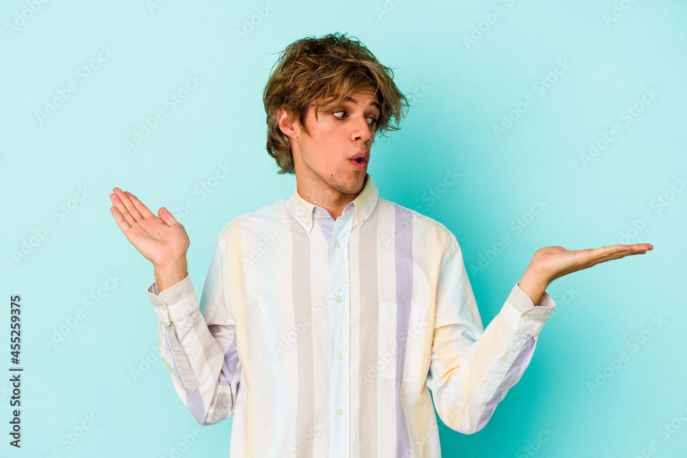 Young caucasian man with make up isolated on blue background confused and doubtful shrugging shoulders to hold a copy space. Young caucasian man with make up isolated on blue background confused and doubtful shrugging shoulders to hold a copy space.