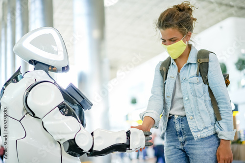 Robot and human handshake in a public place, modern future technological concept. Smart robotic assistant with AI and pretty woman in yellow face mask