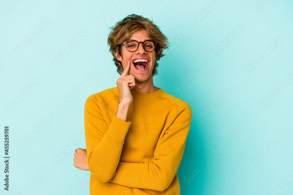 Young caucasian man with make up isolated on blue background smiling happy and confident, touching chin with hand. Young caucasian man with make up isolated on blue background smiling happy and confident, touching chin with hand.