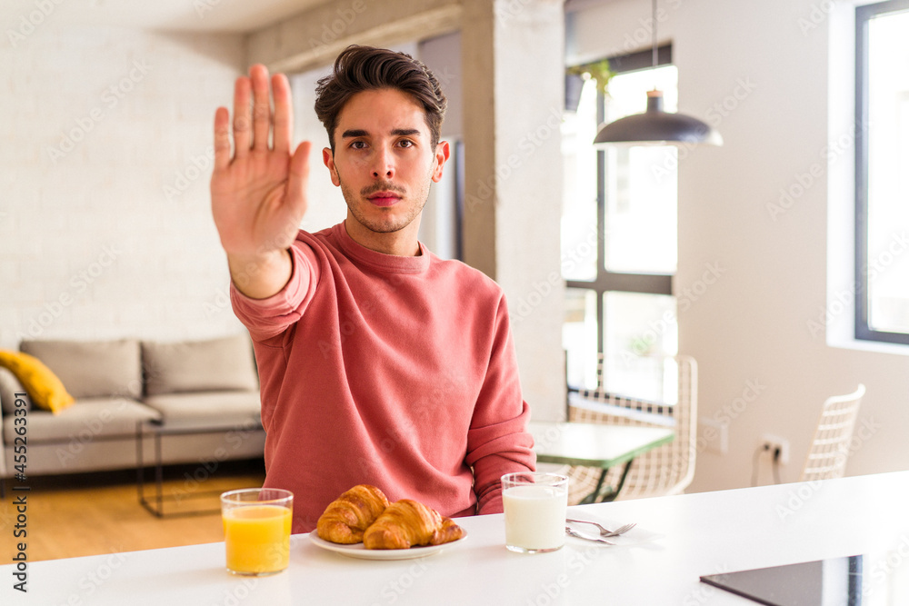 Young mixed race man having breakfast in a kitchen on the morning standing with outstretched hand showing stop sign, preventing you. Young mixed race man having breakfast in a kitchen on the morning standing with outstretched hand showing stop sign, preventing you.