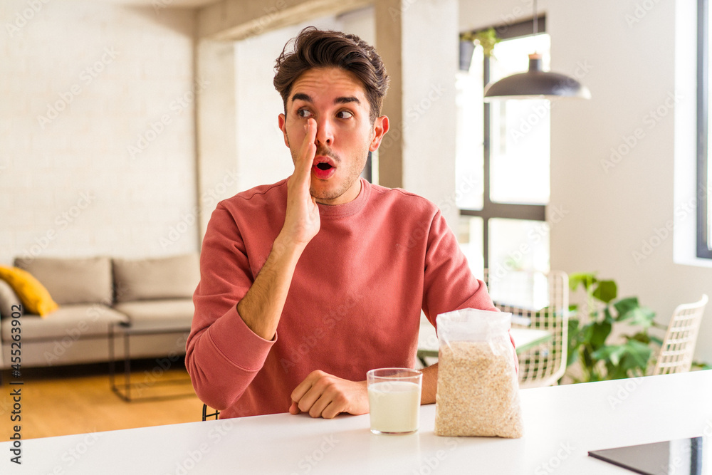 Young mixed race man eating oatmeal and milk for breakfast in his kitchen is saying a secret hot braking news and looking aside Young mixed race man eating oatmeal and milk for breakfast in his kitchen is saying a secret hot braking news and looking aside