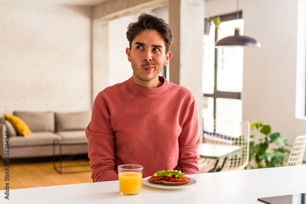 Young mixed race man having breakfast in his kitchen confused, feels doubtful and unsure. Young mixed race man having breakfast in his kitchen confused, feels doubtful and unsure.