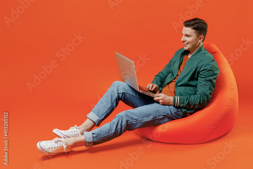Full size body length happy smiling vivid young brunet man 20s wears red t-shirt green jacket sit in bag chair hold use work on laptop pc computer isolated on plain orange background studio portrait