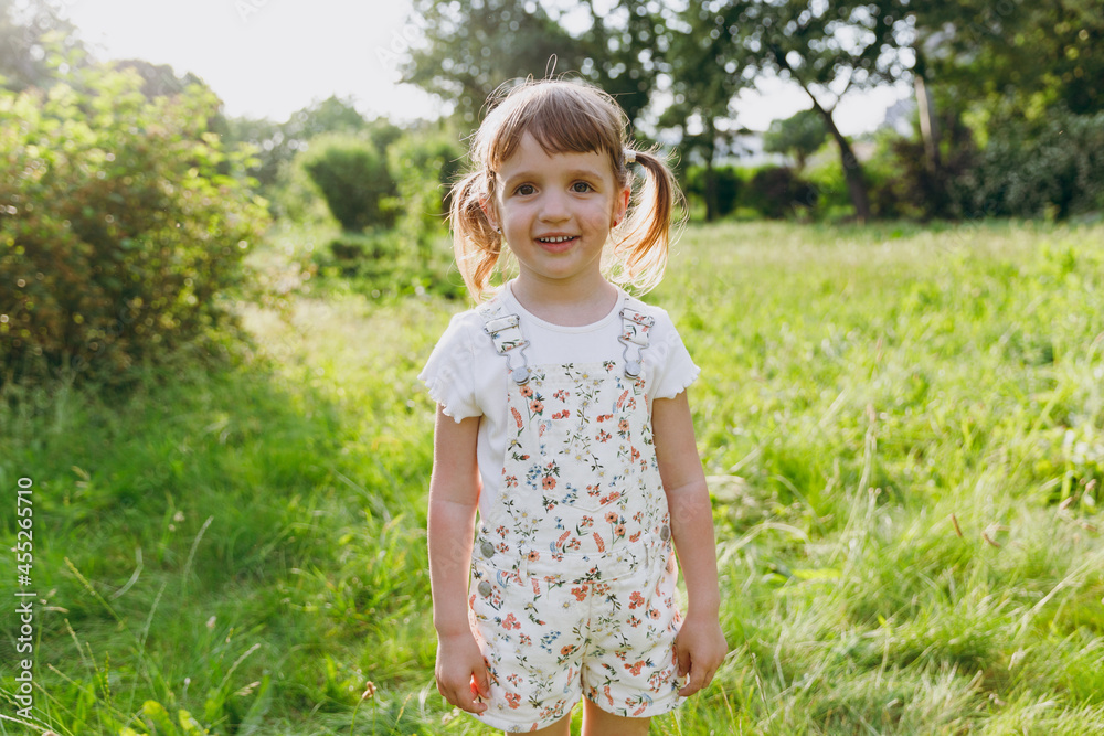 Little smiling happy beautiful kid girl 5-6 years old wear white casual clothes look camera play on park green sunshine lawn, spending time outdoor in village countyside during summer time vacations. Little smiling happy beautiful kid girl 5-6 years old wear white casual clothes look camera play on park green sunshine lawn, spending time outdoor in village countyside during summer time vacations.