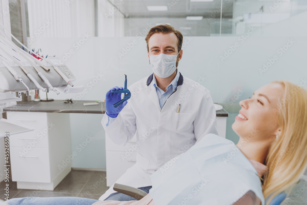 Young european man doctor in face shields and googles hold dental equipment instrument device examine mouth cavity of patient woman sitting at dentist office chair indoor cabinet near stomatologist.