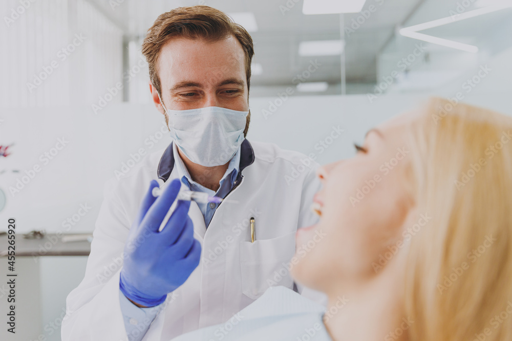 Stomatologist young man doctor in mask hold syringe going to make injection anesthesia for calm patient blonde woman with caries sit at dentist office chair indoor cabinet Healthcare decay treatment. Stomatologist young man doctor in mask hold syringe going to make injection anesthesia for calm patient blonde woman with caries sit at dentist office chair indoor cabinet Healthcare decay treatment.