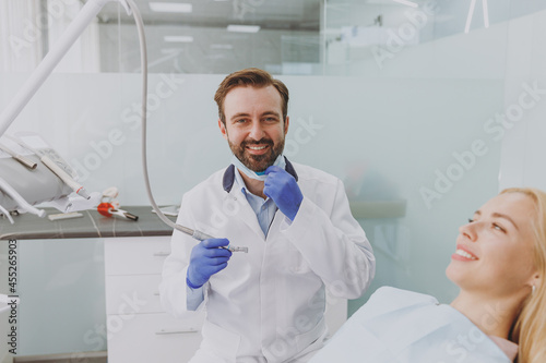 Young man dentist stomatologist doctor wear mask drills tooth of patient caucasian woman with caries decay sit at dentist office chair indoor cabinet near stomatologist. Healthcare enamel treatment