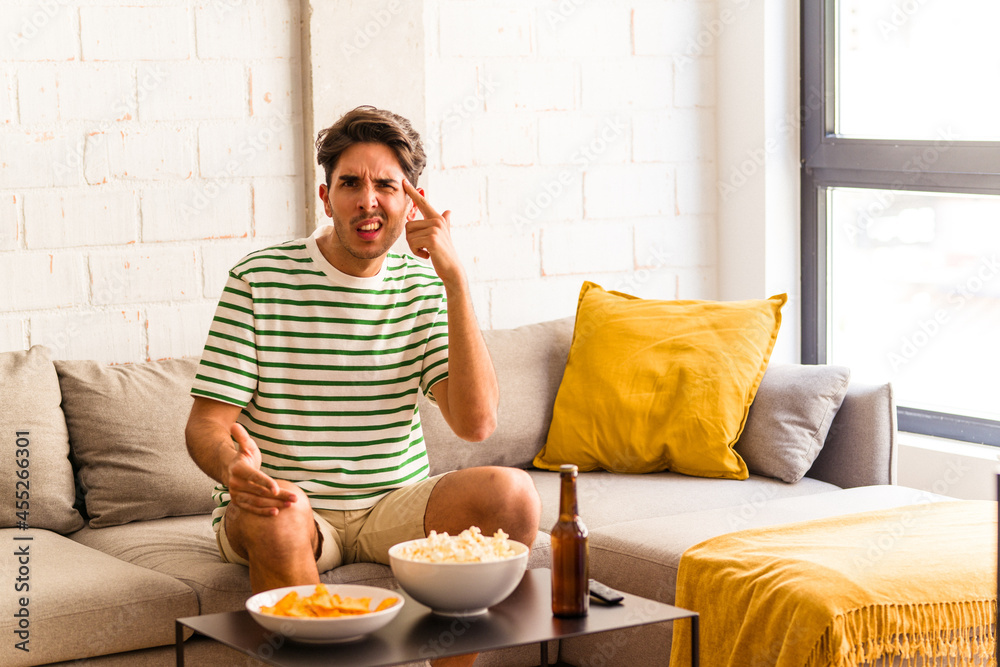 Young mixed race man eating popcorns sitting on the sofa showing a disappointment gesture with forefinger. Young mixed race man eating popcorns sitting on the sofa showing a disappointment gesture with forefinger.