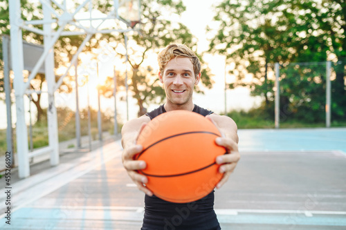 Smiling happy sunlit fun young sporty sportsman man in black sports clothes training look camera holding in hand give ball play at basketball game playground court. Outdoor courtyard sport concept