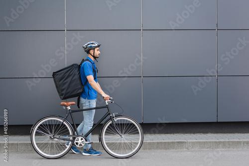 Side view of arabian courier with thermo backpack walking near bike and building outdoors