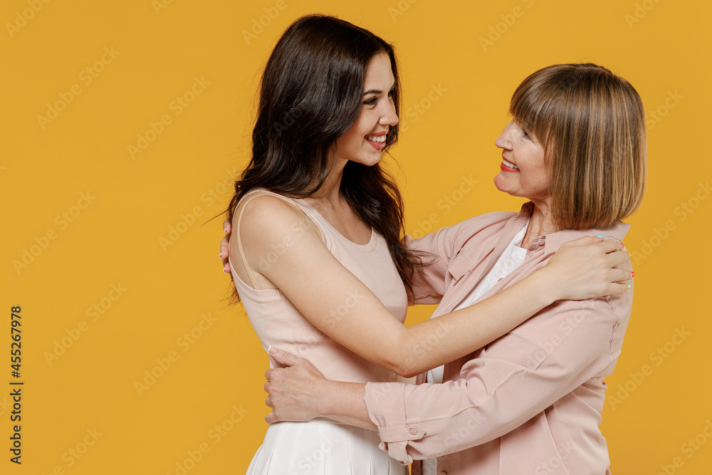 Side view two young smiling happy daughter mother together couple women in casual beige clothes look camera hugging isolated on plain yellow color background studio portrait Family lifestyle concept.