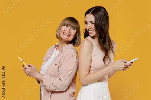 Side view two fun young daughter mother together couple women in casual clothes hold mobile cell phone stand back to back look to each other isolated on plain yellow background studio Family concept.