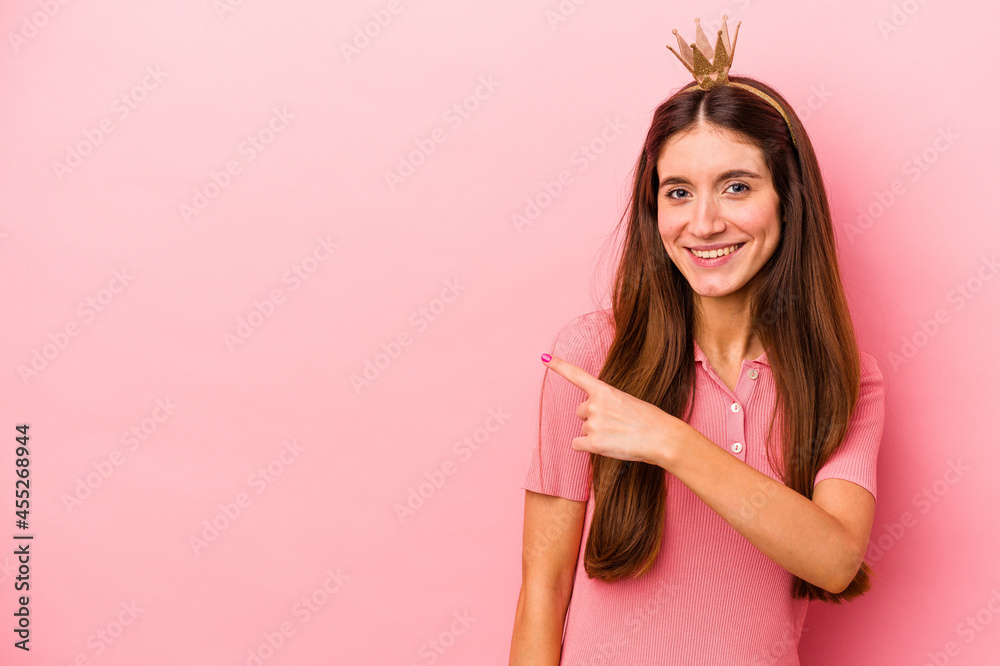 Young caucasian woman wearing crown isolated on pink background smiling and pointing aside, showing something at blank space. Young caucasian woman wearing crown isolated on pink background smiling and pointing aside, showing something at blank space.