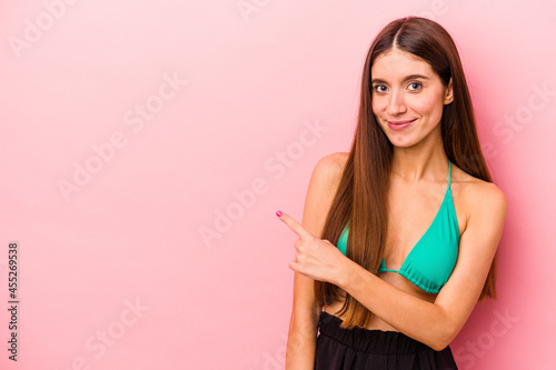 Young caucasian woman wearing bikini isolated on pink background smiling and pointing aside, showing something at blank space.