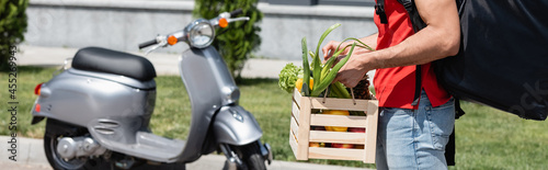 Cropped view of deliveryman with vegetables and backpack standing near blurred scooter outdoors, banner