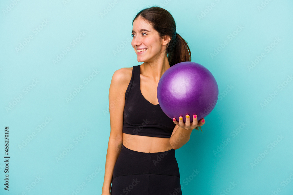 Young caucasian fitness woman holding a ball isolated on blue background looks aside smiling, cheerful and pleasant. Young caucasian fitness woman holding a ball isolated on blue background looks aside smiling, cheerful and pleasant.