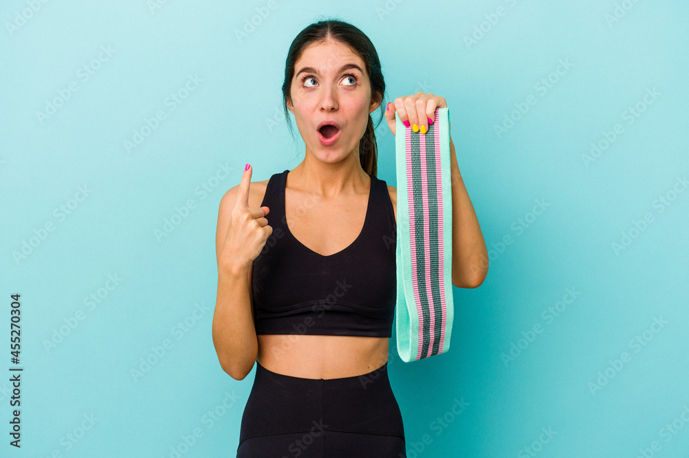 Young caucasian sporty woman holding elastic bands isolated on blue background pointing upside with opened mouth. Young caucasian sporty woman holding elastic bands isolated on blue background pointing upside with opened mouth.