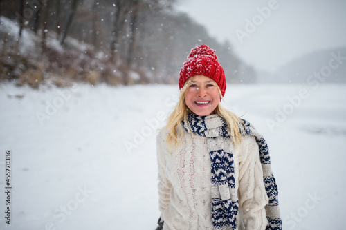 Front view portrait of happy senior woman with hat outdoors standing in snowy nature, looking at camera.