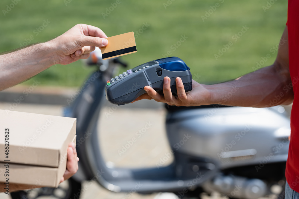 Cropped view of man holding credit card and pizza boxes near courier with terminal near blurred scoter Cropped view of man holding credit card and pizza boxes near courier with terminal near blurred scoter