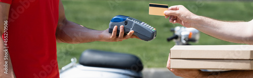 Cropped view of man with pizza boxes holding credit card near courier with payment terminal outdoors, banner