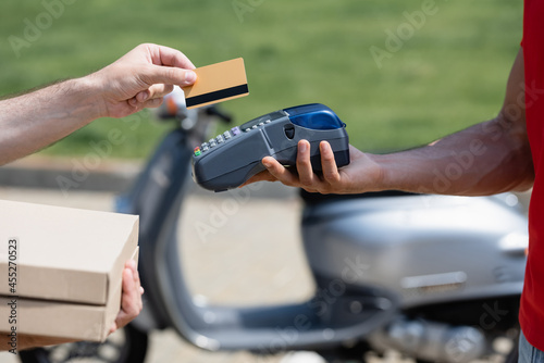 Cropped view of man holding credit card and pizza boxes near courier with terminal near blurred scoter
