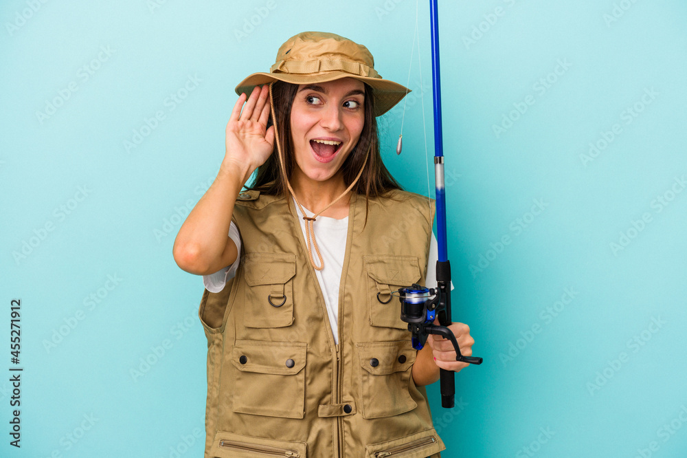 Young caucasian fisherwoman isolated on blue background trying to listening a gossip.