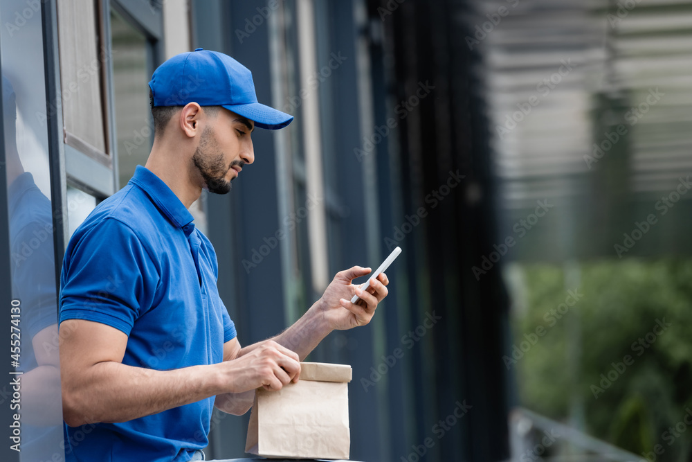 Side view of muslim courier in cap using smartphone and holding paper bag near building Side view of muslim courier in cap using smartphone and holding paper bag near building
