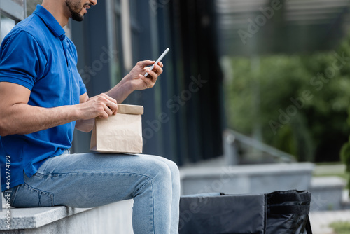 Cropped view of smiling courier using smartphone and holding paper bag outdoors © LIGHTFIELD STUDIOS