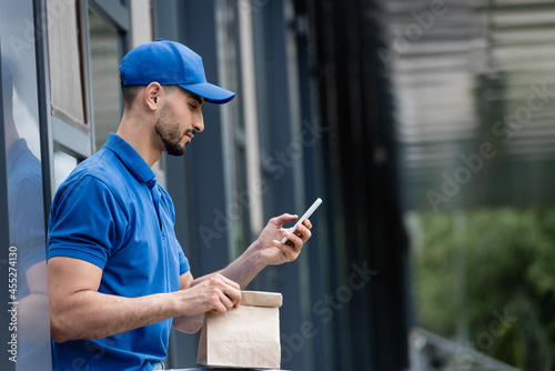 Side view of muslim courier in cap using smartphone and holding paper bag near building © LIGHTFIELD STUDIOS