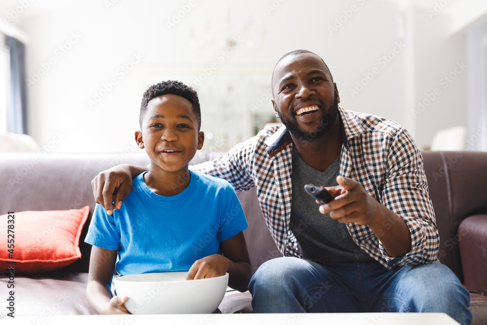 Happy african american father and son sitting on sofa, watching tv