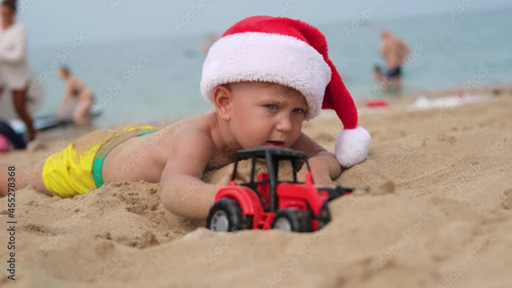 Happy boy in a santa claus hat plays with a toy car on the beach. A child in Christmas is resting on the sea. Copy space Happy boy in a santa claus hat plays with a toy car on the beach. A child in Christmas is resting on the sea. Copy space