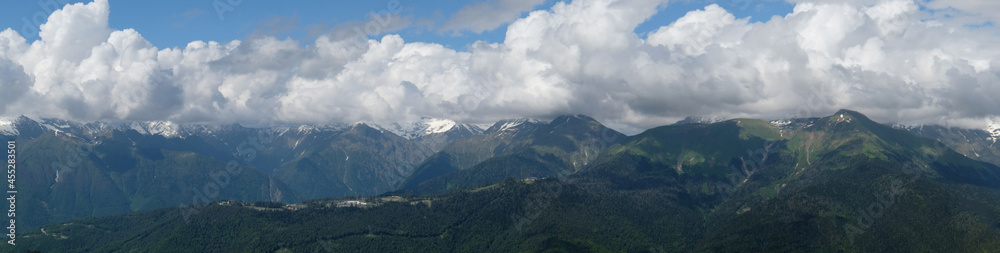 View of green mountains with snowy peaks. Blue sky and clouds cover the mountain peak. Traveling and resting in the mountains in summer, hiking, climbing the mountain. Mountain landscape. View of green mountains with snowy peaks. Blue sky and clouds cover the mountain peak. Traveling and resting in the mountains in summer, hiking, climbing the mountain. Mountain landscape.