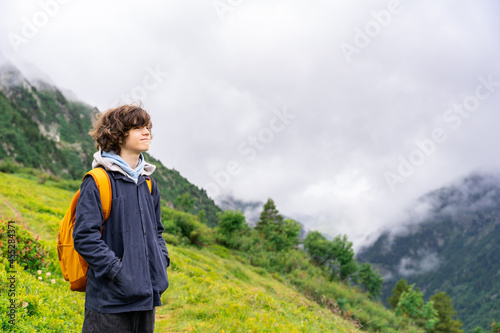 Portrait of a young teenager boy with curly hairs , with yellow backpack in a black jacket standing in the mountains in French Alps, looking away with pleasure. Hiking activities in summer.