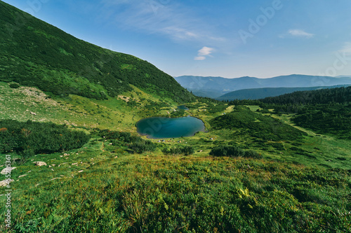 Mountain landscape with forest and lake in sunny summer weather. A camp with tourists' tents is set up near the lake. Climbing to the top, rest, tourism.