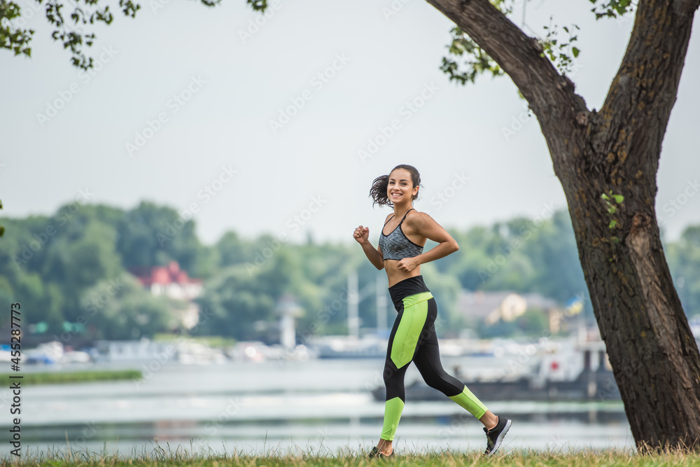 full length of joyful sportswoman in crop top and leggings jogging in park near lake