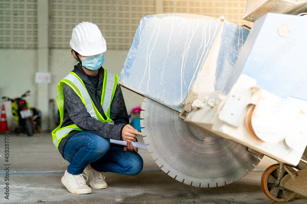 Young Asian female engineer wearing a medical mask to cover her mouth and wearing a white safety helmet Inspecting large floor cutters in the construction zone. Young Asian female engineer wearing a medical mask to cover her mouth and wearing a white safety helmet Inspecting large floor cutters in the construction zone.