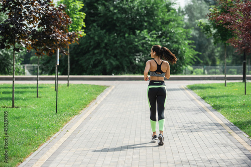 back view of sportive woman in crop top, leggings and wireless earphones listening music while running in park
