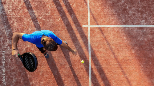 Top view of a paddle tennis player bouncing the ball for a serve on a court.