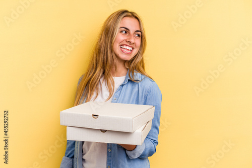 Young caucasian blonde woman holding pizzas isolated on yellow background looks aside smiling, cheerful and pleasant.