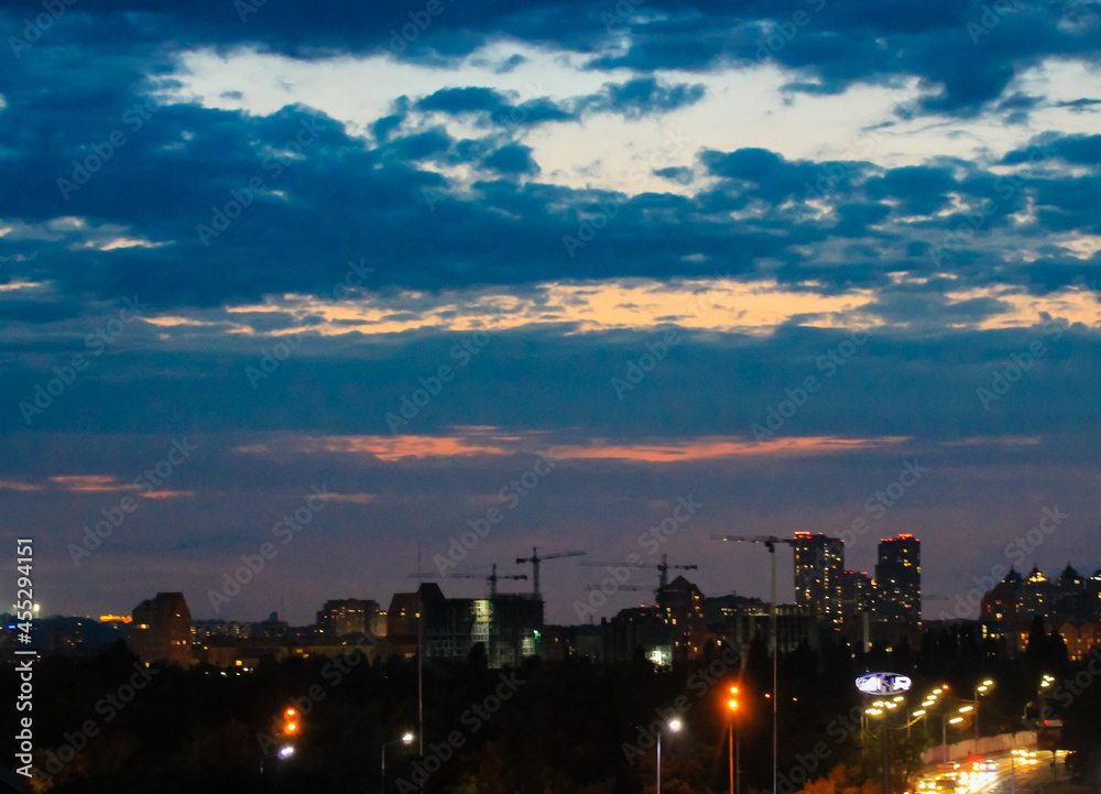 Panorama of a big city at night with a view of high-rise buildings on a horizon. Twilight in a town. Blurred urban background. Lights in the windows, street lamps against dark blue twilight cloudy sky