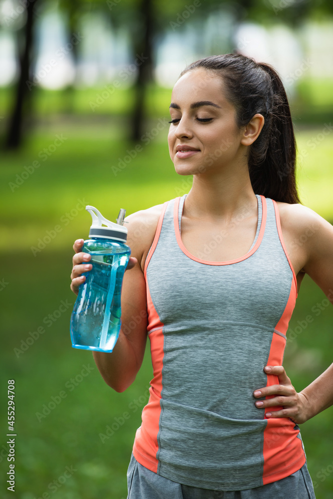 happy and fit woman holding sports bottle while standing with hand on hip happy and fit woman holding sports bottle while standing with hand on hip