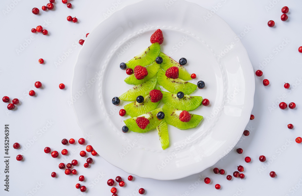 christmas food for kids. white dish with christmas tree made of kiwi, raspberries, cowberry and huckleberry christmas food for kids. white dish with christmas tree made of kiwi, raspberries, cowberry and huckleberry