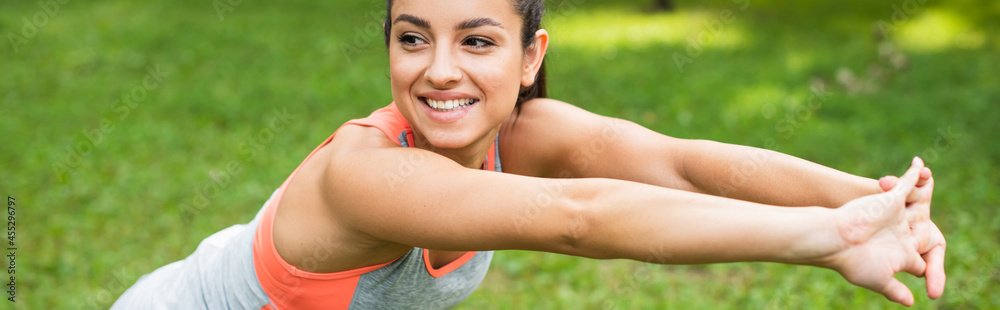 joyful and sportive woman stretching while warming up in park, banner