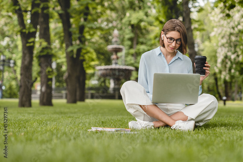 Young caucasian businesswoman student freelancer tutor using laptop for e-learning, distant remote work job occupation, checking e-mails, e-banking online sitting on green grass lawn